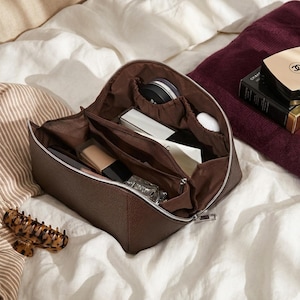 May include: A brown leather cosmetic bag with a silver zip, open to show various makeup products. The bag has multiple compartments and is filled with items like foundation, compacts, and perfume. A tortoiseshell hair clip rests nearby.