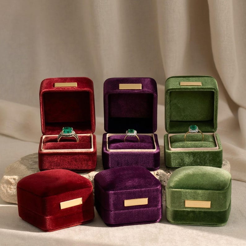 Six premium velvet jewelry boxes in deep burgundy, royal purple, and forest green. Three open cases showcase elegant emerald gemstone rings on plush tan cushions, while three closed boxes highlight minimalist gold metal clasps. Set on a rustic travertine stone with aesthetic shadows, this collection is perfect for wedding flat-lay photography and high-end engagement presentations.