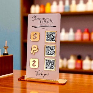 May include: A pink point-of-sale display with the text "Changing Hearts Boutique" and "Thank you!". The display features gold-coloured icons for payment options and QR codes. Makeup brushes and other items are in the background.