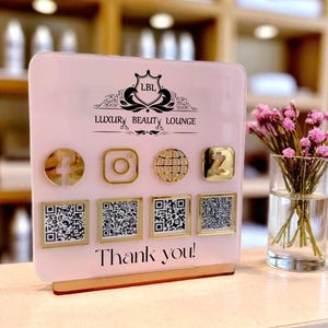 May include: A pink acrylic sign for Luxury Beauty Lounge with gold social media icons and QR codes. The sign includes the text "Thank you!" and a logo. A vase of pink flowers sits to the right.