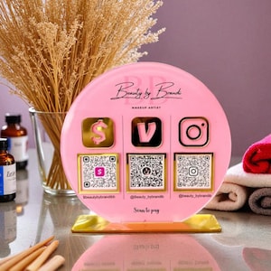 May include: A pink acrylic sign for a makeup artist, featuring social media icons and QR codes. The sign has the text "Beauty by Brandi, Makeup Artist" and "Scan to pay". The sign is on a gold base, with dried flowers in a glass vase in the background.