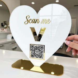 May include: A heart-shaped sign with the words "Scan me" in gold script. The sign has a QR code and a gold "V" on a white background, with a gold base. The sign is designed to be placed on a counter.