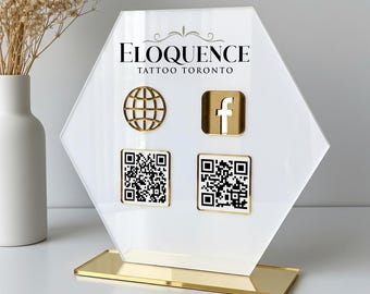 Op de afbeelding: Een wit en goudkleurig acrylbord met de tekst "ELOQUENCE TATTOO TORONTO". Het heeft een gouden wereldbol-icoon, een Facebook-icoon en twee QR-codes. Het bord staat op een gouden voet.