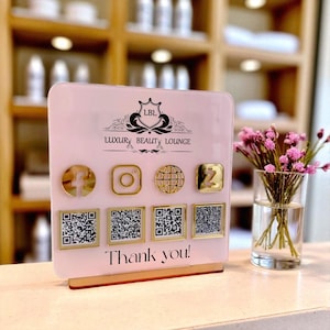 May include: A pink acrylic sign for Luxury Beauty Lounge with gold social media icons and QR codes. The sign includes the text "Thank you!" and a logo. A small vase of pink flowers sits to the right.