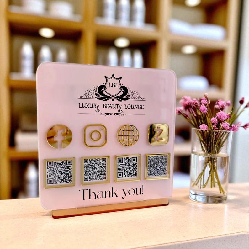 May include: A pink acrylic sign for Luxury Beauty Lounge with gold social media icons and QR codes. The sign includes the text "Thank you!" and a logo. A small vase of pink flowers sits to the right.