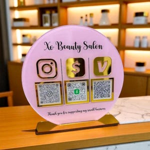 Könnte beinhalten: Ein rosa Acrylschild für einen Schönheitssalon mit Social-Media-Symbolen und QR-Codes. Das Schild hat goldene Akzente und den Text "Xo Beauty Salon". Der Sockel ist goldfarben und das Schild lautet "Vielen Dank für die Unterstützung meines kleinen Unternehmens."