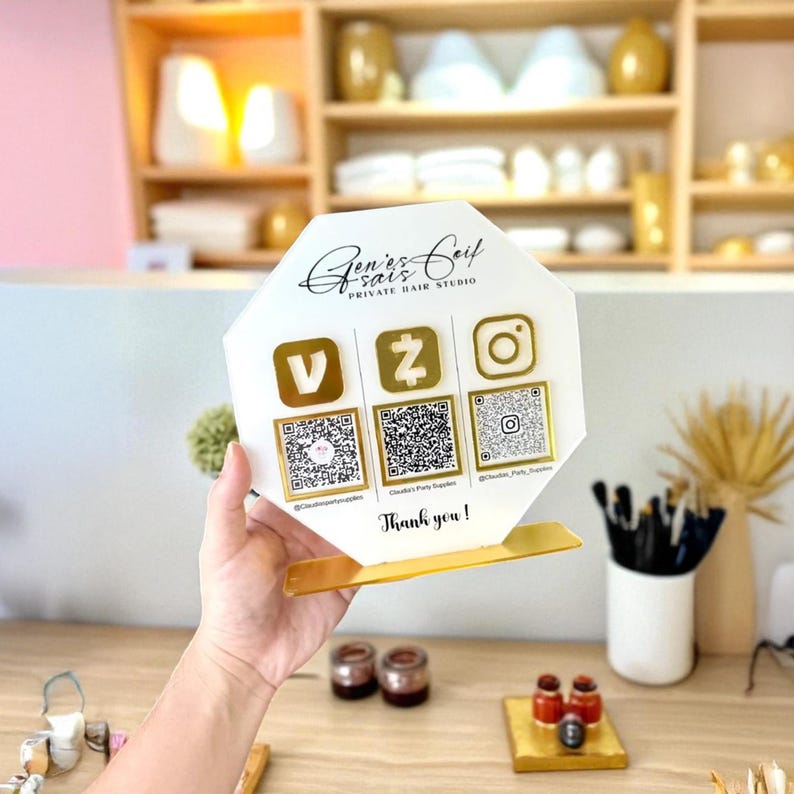 May include: A white and gold sign for Gen's Coif Private Hair Studio, with social media icons and QR codes. The sign has a gold base and the text "Thank you!" at the bottom. The sign is held by a person's hand.