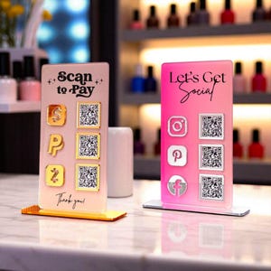Peut inclure: Deux présentoirs en acrylique pour les paiements et les réseaux sociaux. L'un est rose avec le texte "Let's Get Social" et des icônes de réseaux sociaux. L'autre est rose clair avec le texte "Scan to Pay" et des icônes de paiement. Les deux ont des codes QR.