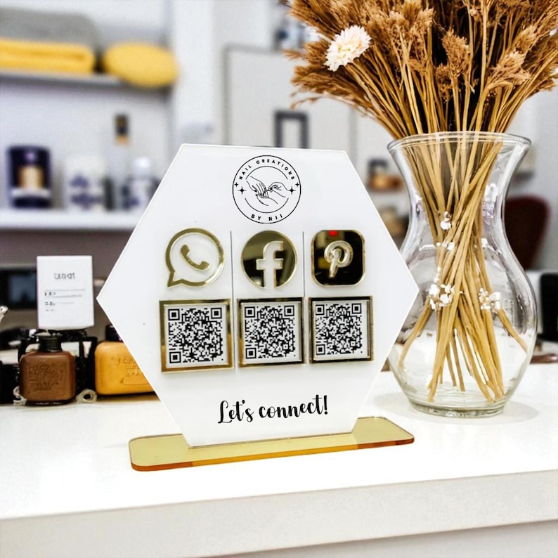 May include: A white hexagonal display with gold detailing showcases social media icons and QR codes for WhatsApp, Facebook, and Pinterest. The text "Let's connect!" is displayed. A vase of dried flowers is to the right.
