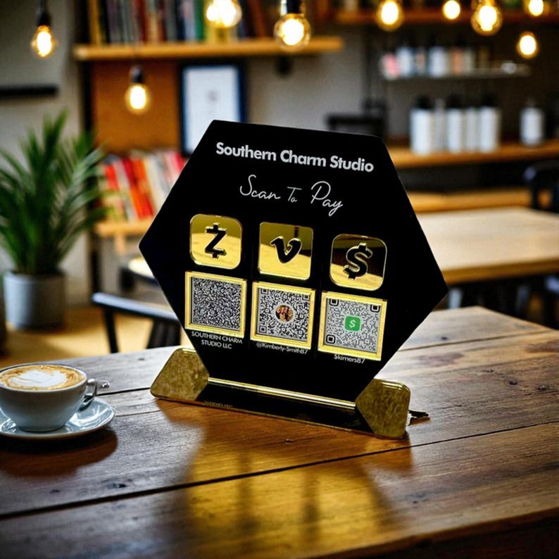 May include: A black and gold payment display with the text "Southern Charm Studio" and "Scan To Pay". The display features QR codes and payment symbols. A cup of coffee sits on a wooden table.