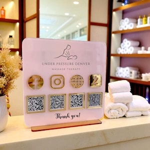 May include: A pink sign for "Under Pressure Denver Massage Therapy" with gold social media icons and QR codes. The sign says "Thank you!" at the bottom. White towels are stacked on a counter.