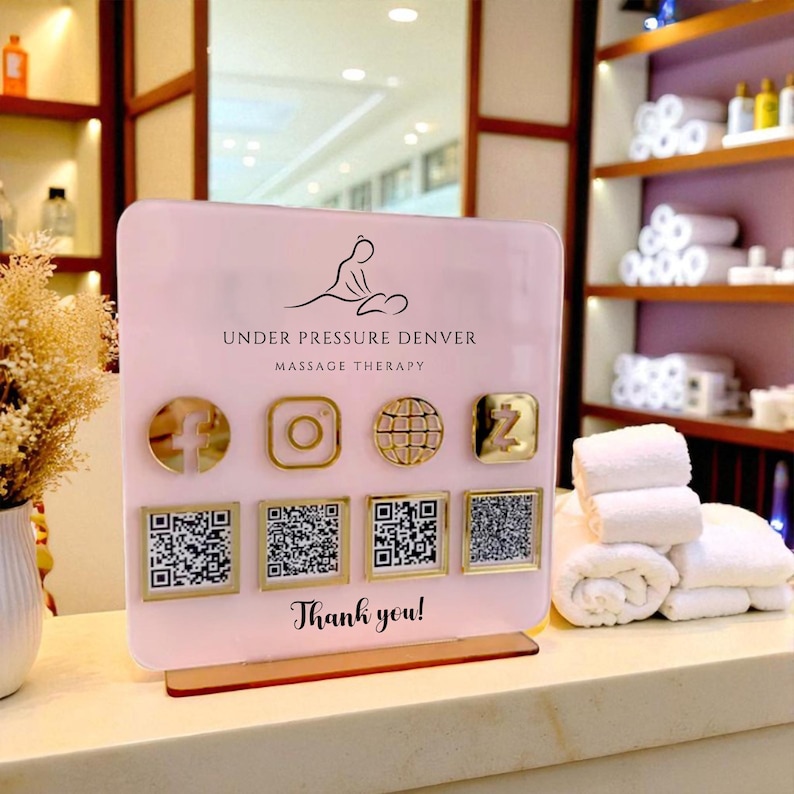 May include: A pink sign for "Under Pressure Denver Massage Therapy" with gold social media icons and QR codes. The sign says "Thank you!" at the bottom. White towels are stacked on a counter.