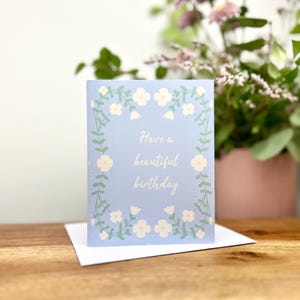 May include: A light blue rectangular greeting card featuring a white floral vine border and the text Have a beautiful birthday printed in white in the center. The card is positioned on a white envelope on a wooden surface.