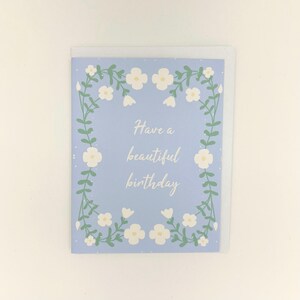 May include: A rectangular greeting card with a light blue background featuring a white floral border of vines and small flowers. The center of the card displays the white cursive text Have a beautiful birthday. The card is paired with a white envelope.