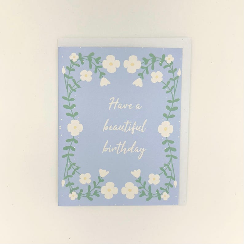 May include: A rectangular greeting card with a light blue background featuring a white floral border of vines and small flowers. The center of the card displays the white cursive text Have a beautiful birthday. The card is paired with a white envelope.