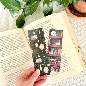 May include: Two decorative bookmarks with dark green backgrounds. One features books, potion bottles, and floral accents. The other depicts a bookshelf with books, a skull, and other items. The bookmarks are held over an open book.