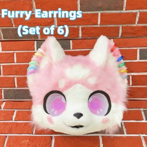 May include: Furry earrings shaped like a cartoon animal face, featuring pink, white, and black details. The earrings have large eyes and are decorated with colourful bands. The text "Furry Earrings (Set of 6)" is displayed above the earrings.