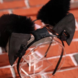 May include: Black cat ear headband with fluffy black ears and a clear plastic headband. The headband has black elastic straps that attach to the ears.