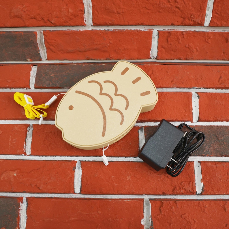 Puede incluir: Una l&aacute;mpara LED beige con forma de pez y detalles marrones, montada en una pared de ladrillos rojos. Incluye un cable de alimentaci&oacute;n amarillo y un adaptador de corriente negro. La l&aacute;mpara tiene un dise&ntilde;o decorativo.