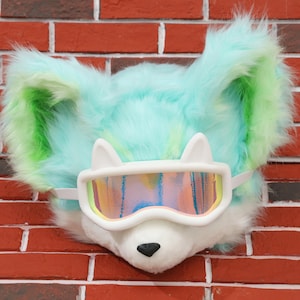 May include: A plush, furry headpiece in shades of blue and green, resembling a fox or similar animal. It features white goggles with a rainbow-colored lens and a white elastic strap. The headpiece has a black nose and is set against a brick wall.
