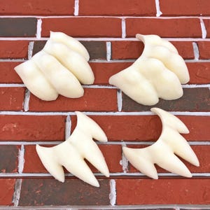May include: Four off-white, claw-shaped objects are arranged against a red brick wall. The objects have a smooth, rounded appearance, with three or four distinct points or fingers extending from a central base. The brick wall has white mortar lines.