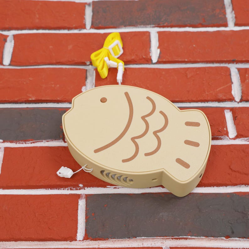 Taiyaki Fish Shaped Portable Fan held by a hand, illustrating its perfect fit for easy portability and usage. The fan’s fun and cute Japanese street snack-inspired design makes it a must-have for anime and kawaii fans, and the 3D printed PLA construction ensures it’s sturdy and environmentally friendly. Whether you’re headed to a summer festival, gaming marathon, or vacation getaway, this USB rechargeable fan delivers a three-speed breeze to keep you comfortable and cool.