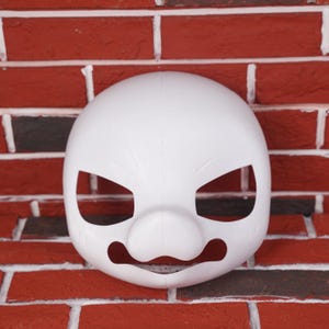 Könnte beinhalten: Eine weiße Maske mit ausgeschnittenen Augen- und Mundöffnungen. Die Maske hat eine abgerundete Form mit einer markanten Nase und leicht gerunzelten Augenbrauen. Die Maske ist vor einer roten Backsteinmauer platziert.