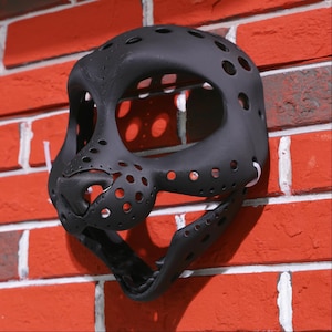 May include: A black plastic mask with a detailed design and multiple holes. The mask is designed to cover the face and has a large opening for the eyes.