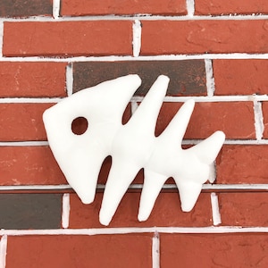 May include: A white, plush fish skeleton decoration against a red brick wall. The fish bone design has a circular eye socket and multiple pointed bone extensions. The background is a classic red brick pattern.
