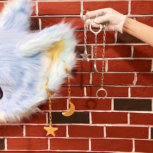 May include: Gold star and moon earrings with chain detailing, and a set of silver celestial-themed earrings, are shown against a red brick wall. A furry blue and white ear is visible on the left side of the image.