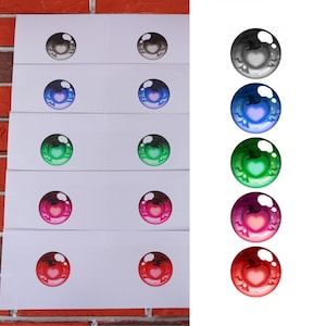 May include: A collection of colorful, cartoon-style eye designs. Each eye features a heart-shaped pupil and is available in various colors, including gray, blue, green, pink, and red. The eyes are displayed on white rectangular cards.