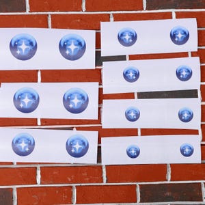 May include: A set of eight white rectangular stickers with a blue star design. Each sticker features a blue circle with a white star in the center. The stickers are arranged in rows on a brick wall.