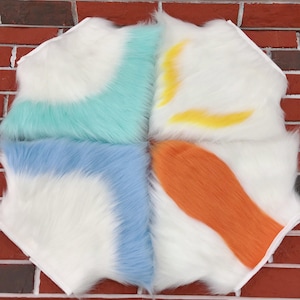 May include: A multi-colored faux fur square with white, blue, teal, yellow, and orange sections. The fur is long and fluffy, with each color section forming a unique shape. The square is set against a brick wall.