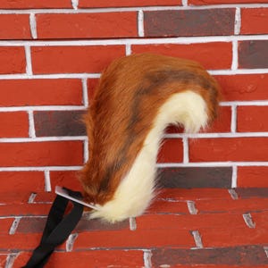 Op de afbeelding: Een bruin-witte nepbontstaart met een zwarte riem en gesp. De staart is een mix van bruin en wit bont, met een donzige textuur. De riem is gemaakt van zwarte stof en is bevestigd aan een doorzichtig plastic stuk.