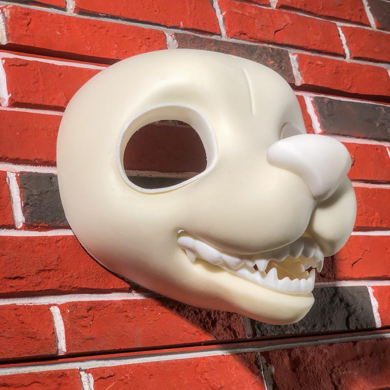 May include: A white unfinished furry animal mask with a large open mouth and teeth. The mask has a large eye hole and a small nose.
