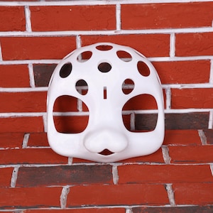 May include: A white mask with large eye and mouth openings, and several circular holes across the top. The mask has a rounded shape and is set against a red brick wall.