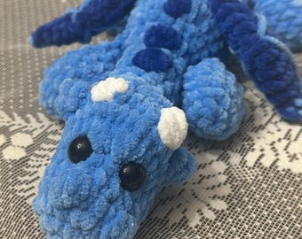 May include: A blue crocheted dragon toy with white accents and black eyes. The dragon is lying on a white crocheted surface.