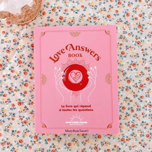 Op de afbeelding: Een roze boek met de titel "Love Answers Book" in rode letters. Het boek toont een rood hart in een kristallen bol die door twee handen wordt vastgehouden. De tekst "Le livre qui répond à toutes tes questions" is in het Frans. Het boek ligt op een oppervlak met een bloemenpatroon.