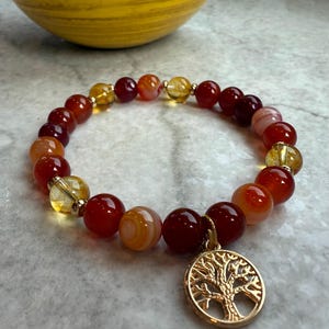 May include: A beaded bracelet featuring a mix of red, orange, and yellow gemstone beads. The bracelet has a gold-colored tree of life charm. The beads are round and vary in size. The bracelet is on a light gray surface.
