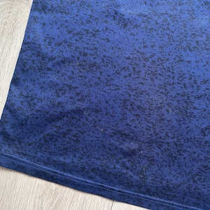 May include: Close-up of a blue fabric with a speckled pattern. The fabric has a dark blue base color with a lighter blue gradient and black speckles. The edge of the fabric is visible.
