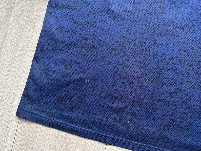 May include: Close-up of a blue fabric with a speckled pattern. The fabric has a dark blue base color with a lighter blue gradient and black speckles. The edge of the fabric is visible.