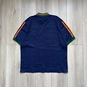 May include: A navy blue short-sleeved shirt with orange stripes on the sleeves and a green and orange collar. The shirt has a textured pattern and is viewed from the back.