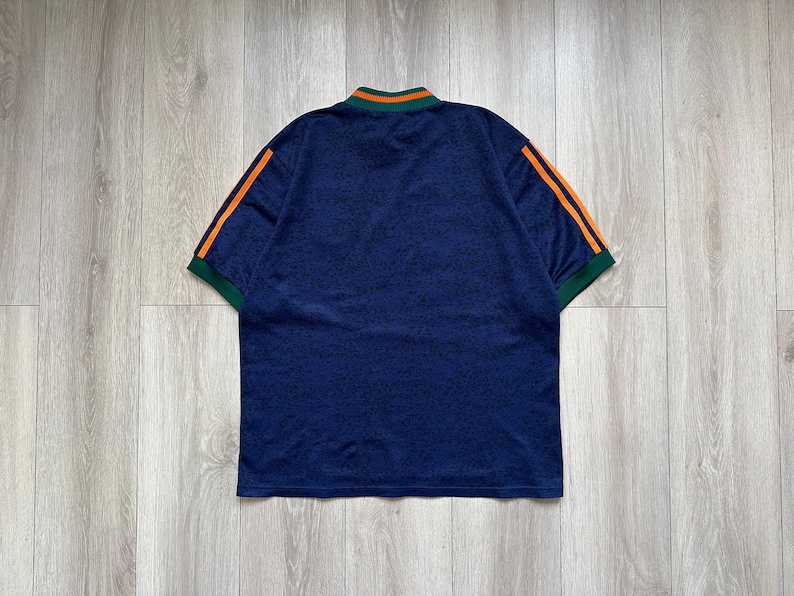 May include: A navy blue short-sleeved shirt with orange stripes on the sleeves and a green and orange collar. The shirt has a textured pattern and is viewed from the back.