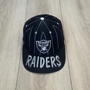 Puede incluir: Gorra de béisbol de los Raiders en blanco y negro. La gorra presenta rayas blancas, el logotipo de los Raiders en la parte delantera y la palabra "RAIDERS" bordada en la visera. Tiene una visera plana y un botón en la parte superior.