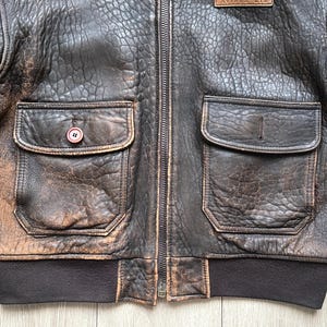 May include: A dark brown distressed leather bomber jacket featuring a full-length front zip, two large front pockets with flaps, and a dark brown ribbed knit waistband. One pocket has a single button closure, while the other is plain.