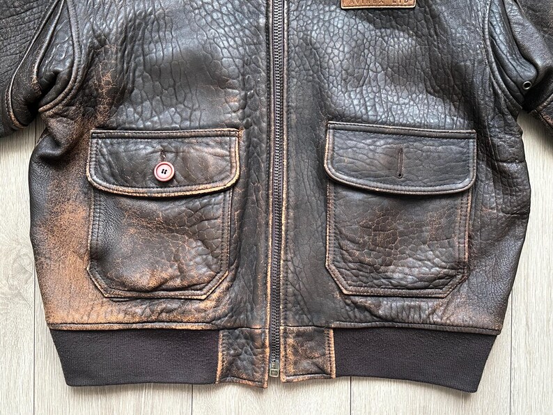 May include: A dark brown distressed leather bomber jacket featuring a full-length front zip, two large front pockets with flaps, and a dark brown ribbed knit waistband. One pocket has a single button closure, while the other is plain.