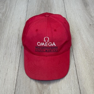 Puede incluir: Gorra de béisbol roja con el texto blanco "OMEGA" y el símbolo Omega. Debajo del nombre de la marca, el texto "OMEGA EUROPEAN MASTERS CRANS MONTANA" está impreso en blanco. La gorra es de material suave.