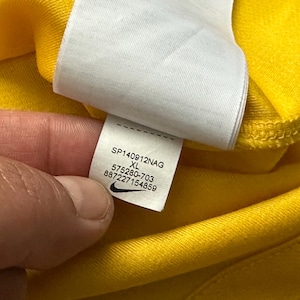 Pu&ograve; includere: Primo piano di un capo giallo con un'etichetta bianca. L'etichetta mostra il testo "SP140912NAG XL 575280-703 887227154859" e un logo Nike. Il capo &egrave; realizzato in un tessuto morbido, possibilmente sportivo.