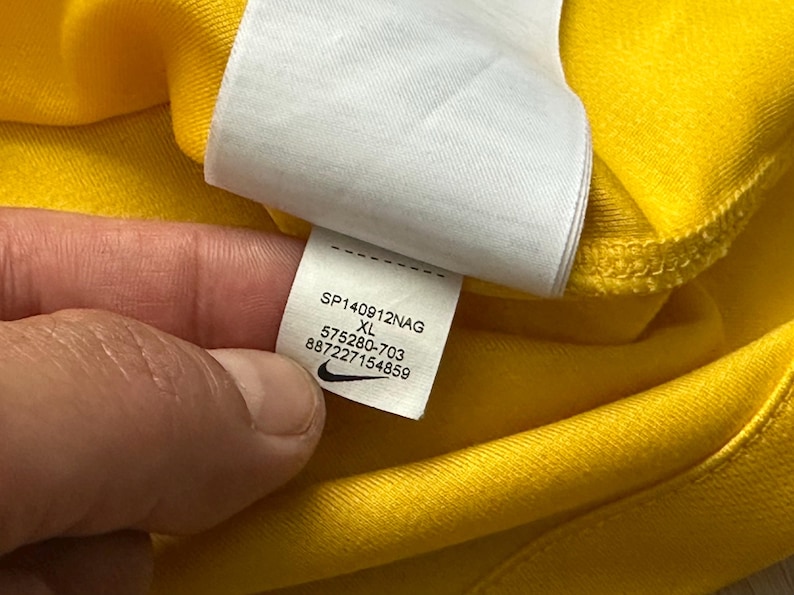 Pu&ograve; includere: Primo piano di un capo giallo con un'etichetta bianca. L'etichetta mostra il testo "SP140912NAG XL 575280-703 887227154859" e un logo Nike. Il capo &egrave; realizzato in un tessuto morbido, possibilmente sportivo.