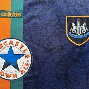 May include: Close-up of a navy blue soccer jersey with orange and green stripes. Features the Adidas logo, a Newcastle United crest, and a Newcastle Brown Ale patch.